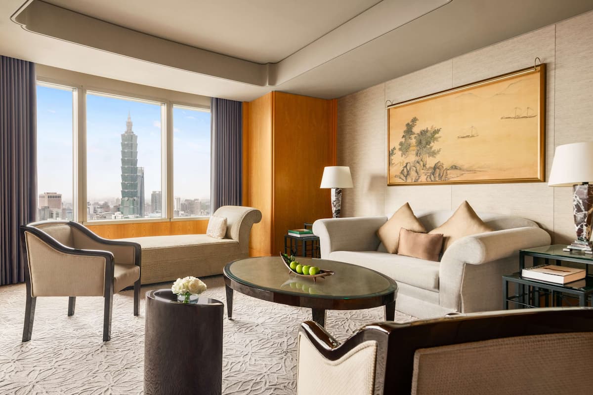 A modern hotel room with a large window offering a city view, a sofa, a chair, a coffee table, and a painting on the wall.