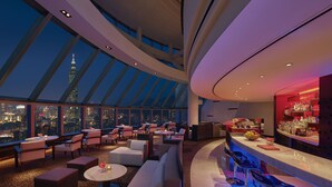Un hall d’hôtel moderne avec un plafond haut, de grandes fenêtres donnant sur la ville et un espace bar avec des sièges.