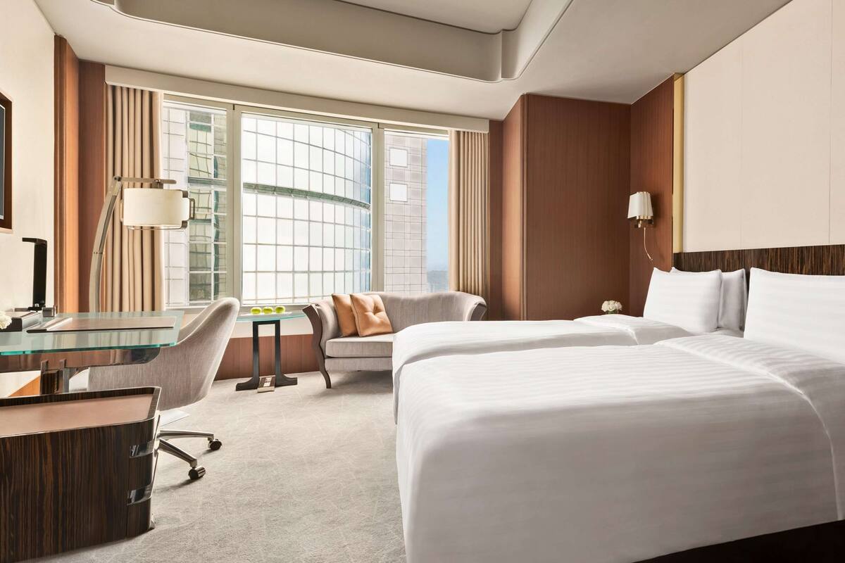 A hotel room with a large bed, a desk with a chair, a small table with a vase of flowers, and a large window with a view of buildings.