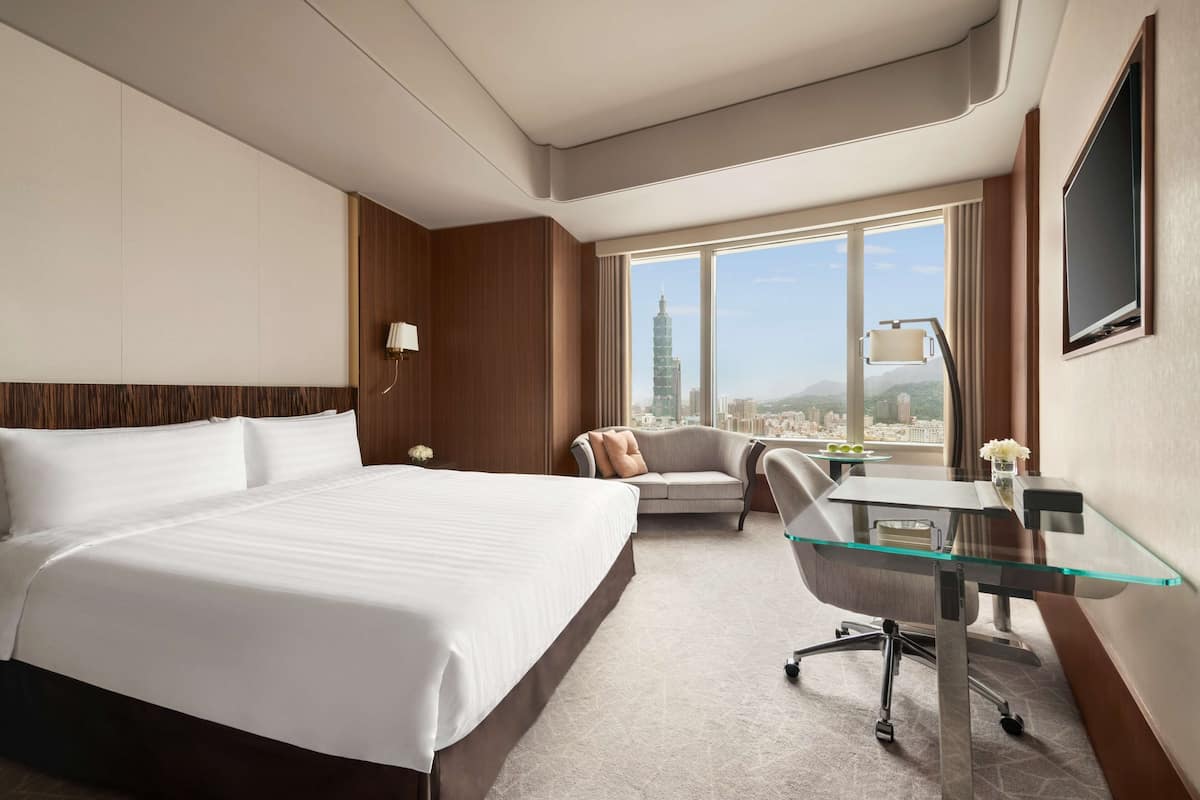 A hotel room with a large bed, a desk with a chair, a sofa, and a large window offering a city view.