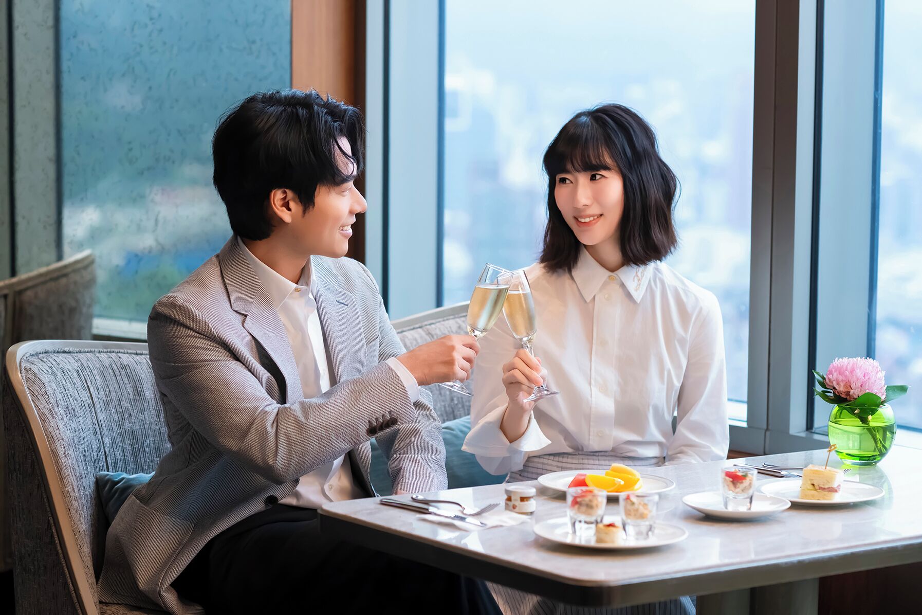 A dining table with a variety of dishes, a green vase with a flower, and a city view through large windows.