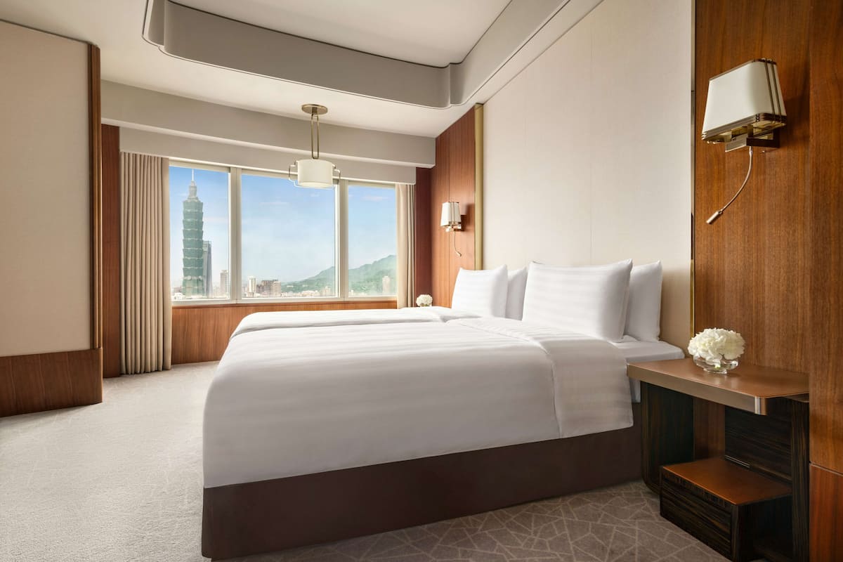 A hotel room with a large bed, a nightstand, a lamp, and a view of a cityscape.