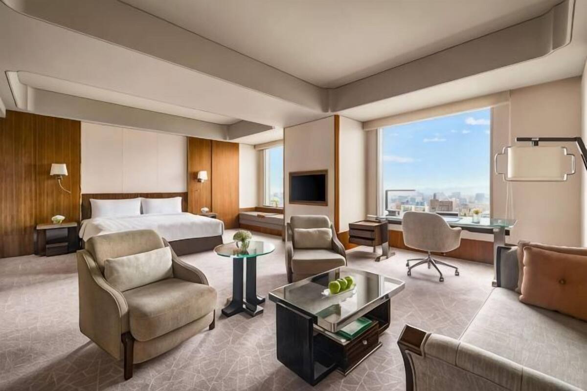A hotel room with a bed, two armchairs, a coffee table, a TV, and a city view.
