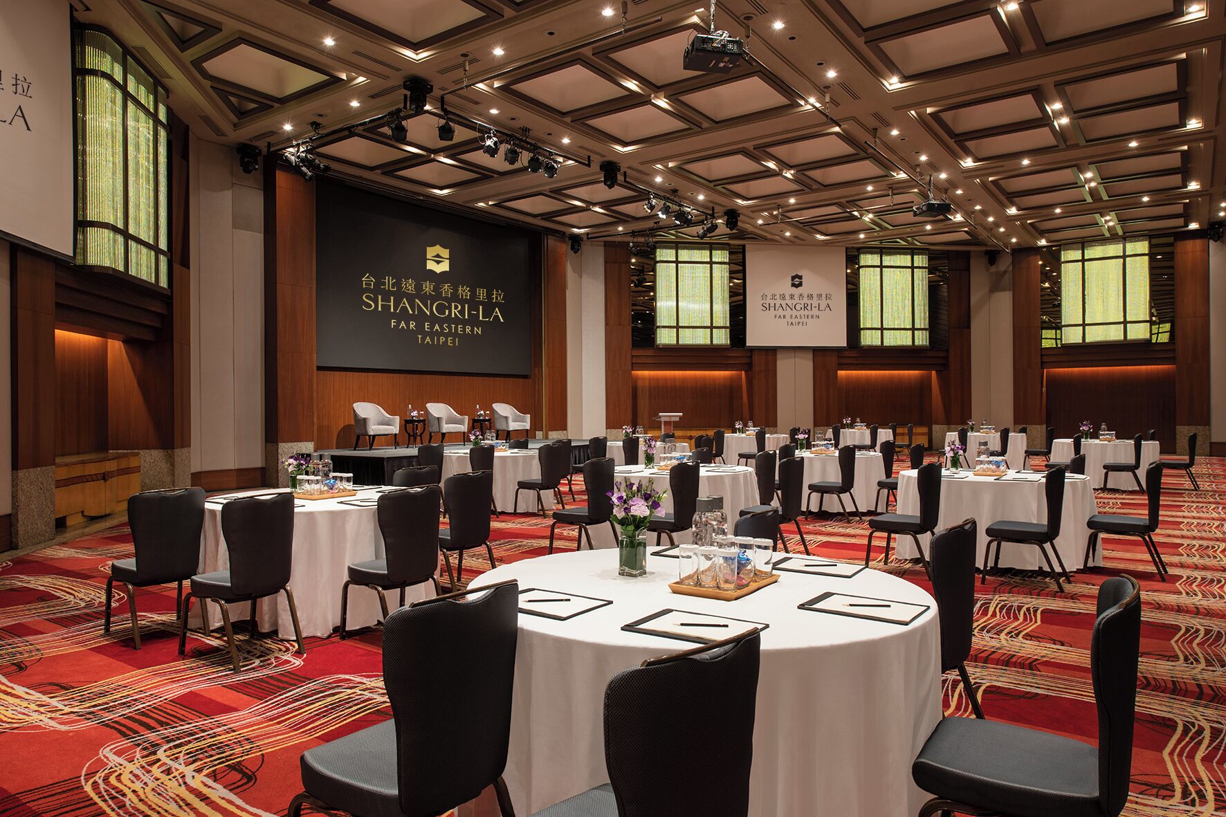 Een congreszaal met ronde tafels, zwarte stoelen en een podium waarop een scherm staat met 'SHANGRI-LA FAR EASTERN TAIPEI'.