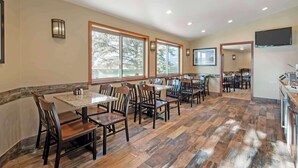 A spacious dining area with multiple tables and chairs, large windows with a view of trees, and a flat-screen TV mounted on the wall. - Best Western Ptarmigan Lodge (Dillon)