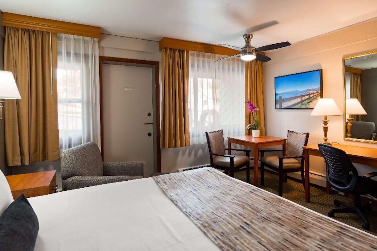 A hotel room with a bed, a desk, two chairs, a desk lamp, a ceiling fan, and a large mirror.