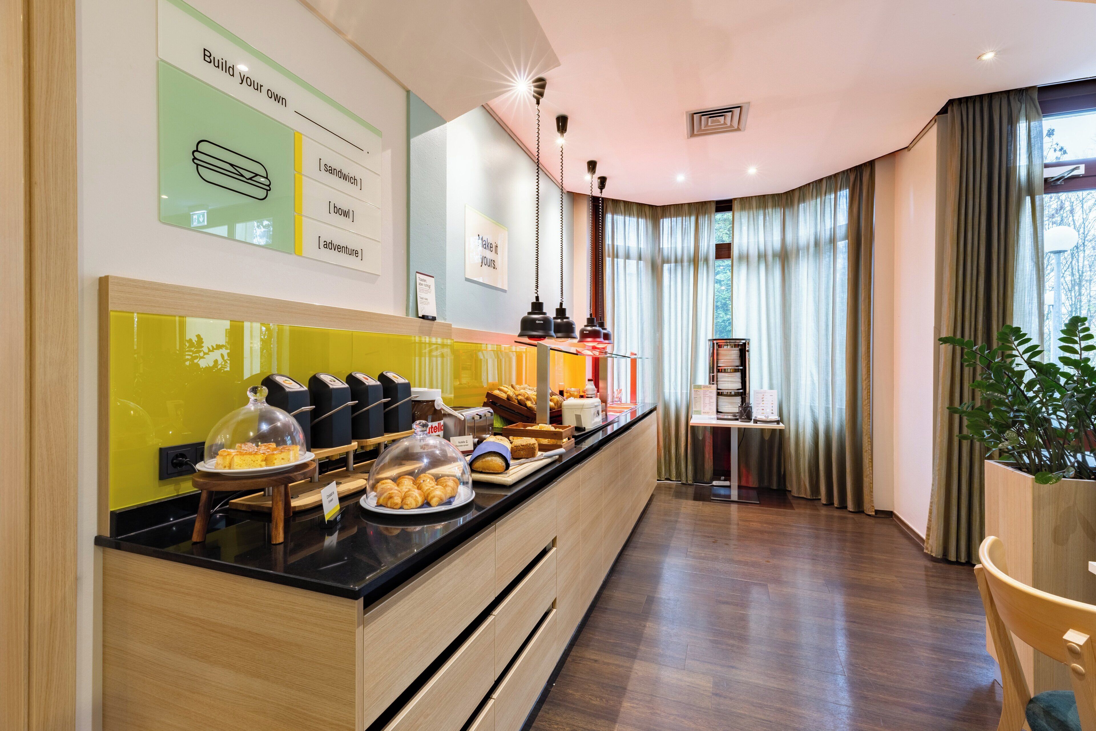 A hotel reception area with a counter displaying breakfast items, a sign with a sandwich illustration, and a view of the outdoors through large windows.