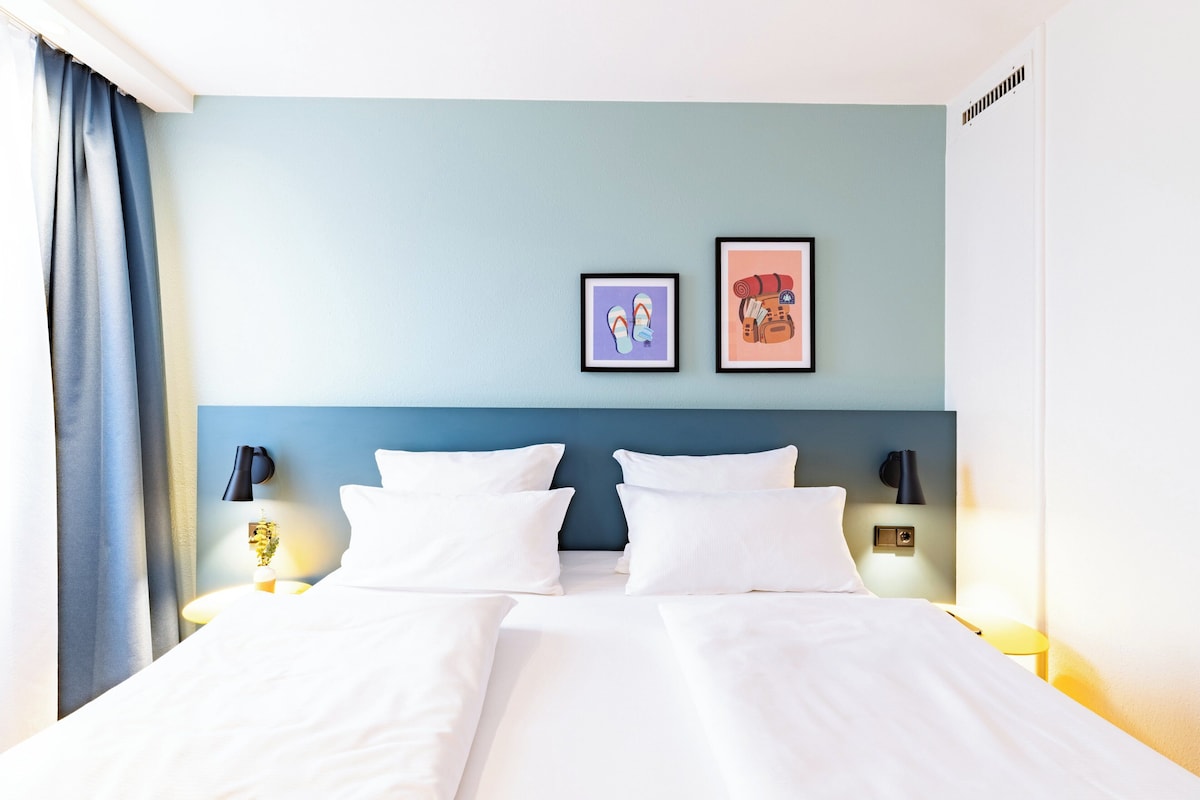 A hotel room with a bed, two wall-mounted lamps, a blue headboard, and two framed pictures on the wall.