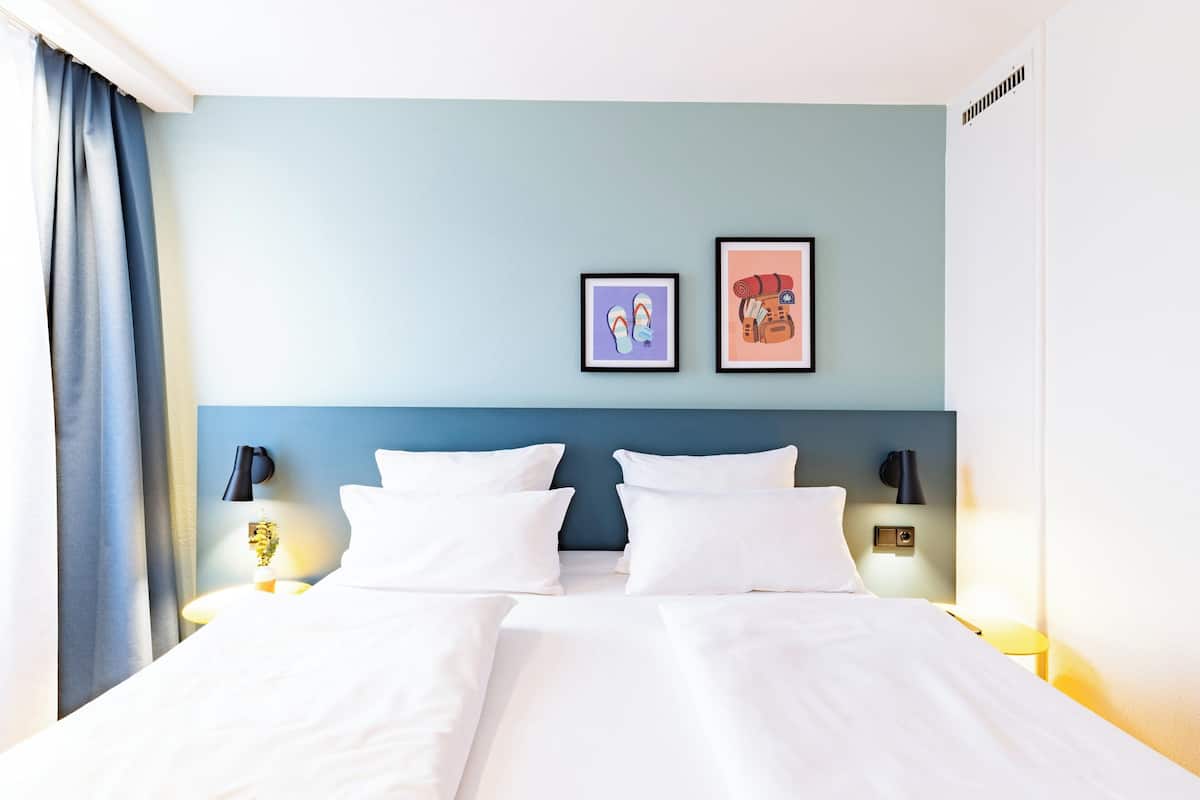 Ett hotellrum med en säng, två väggmonterade lampor, en blå sänggavel och två inramade tavlor på väggen.