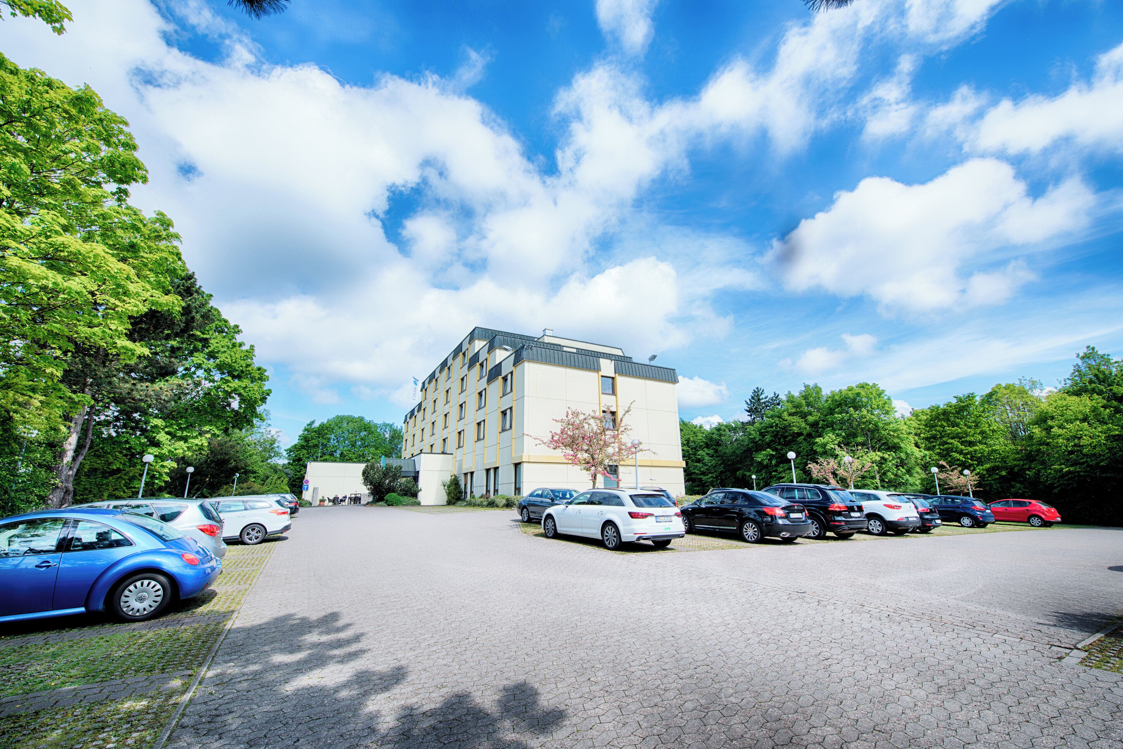 Un parking où plusieurs voitures sont garées devant un bâtiment doté d’un toit plat et d’une façade beige.