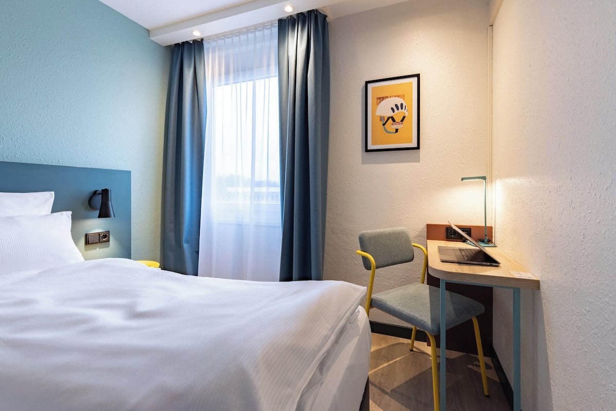Ett hotellrum med en säng, ett skrivbord med en bärbar dator, en stol, ett nattduksbord, en lampa och ett fönster med gardiner.