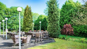 An outdoor patio area with wicker furniture and street lamps, surrounded by lush greenery and trees. - Garner Hotel Osnabruck by IHG (Osnabrueck)