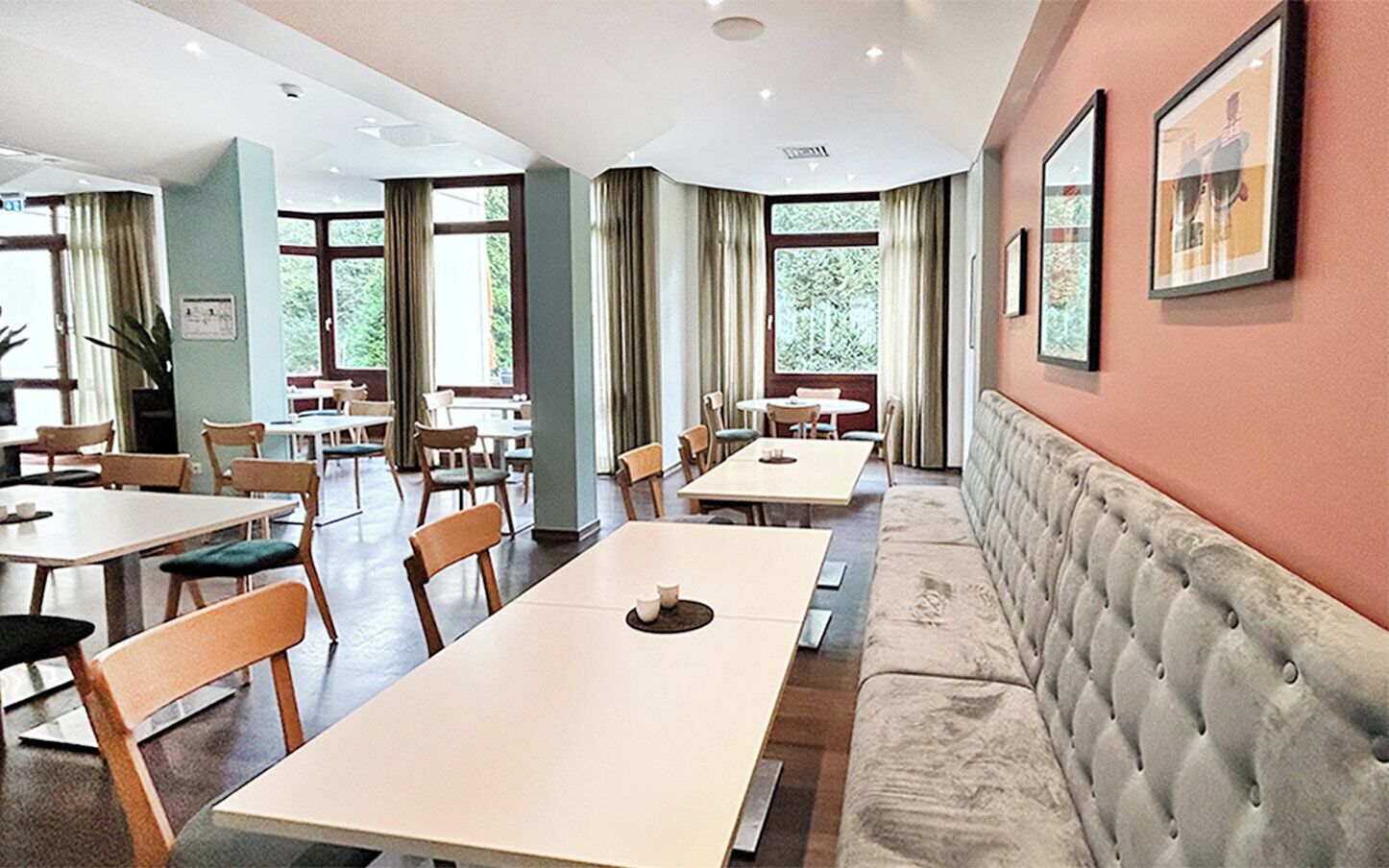 A spacious dining area with multiple tables and chairs, large windows with curtains, and a cushioned bench along the wall.