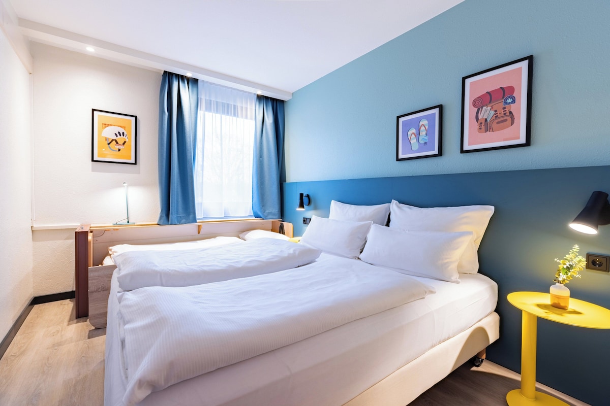 A hotel room with a large bed, a bedside table, a lamp, and framed artwork on the wall.