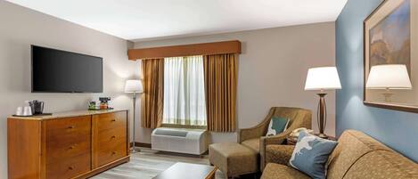 A hotel room with a flat-screen TV, a wooden dresser, a sofa, a coffee table, and a window with curtains.