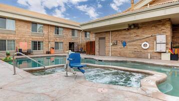 A pool area with a hot tub, a lounge chair, and a brick building with windows and doors.