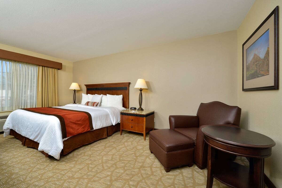 A hotel room with a large bed, a chair, a small table, and a nightstand with lamps.