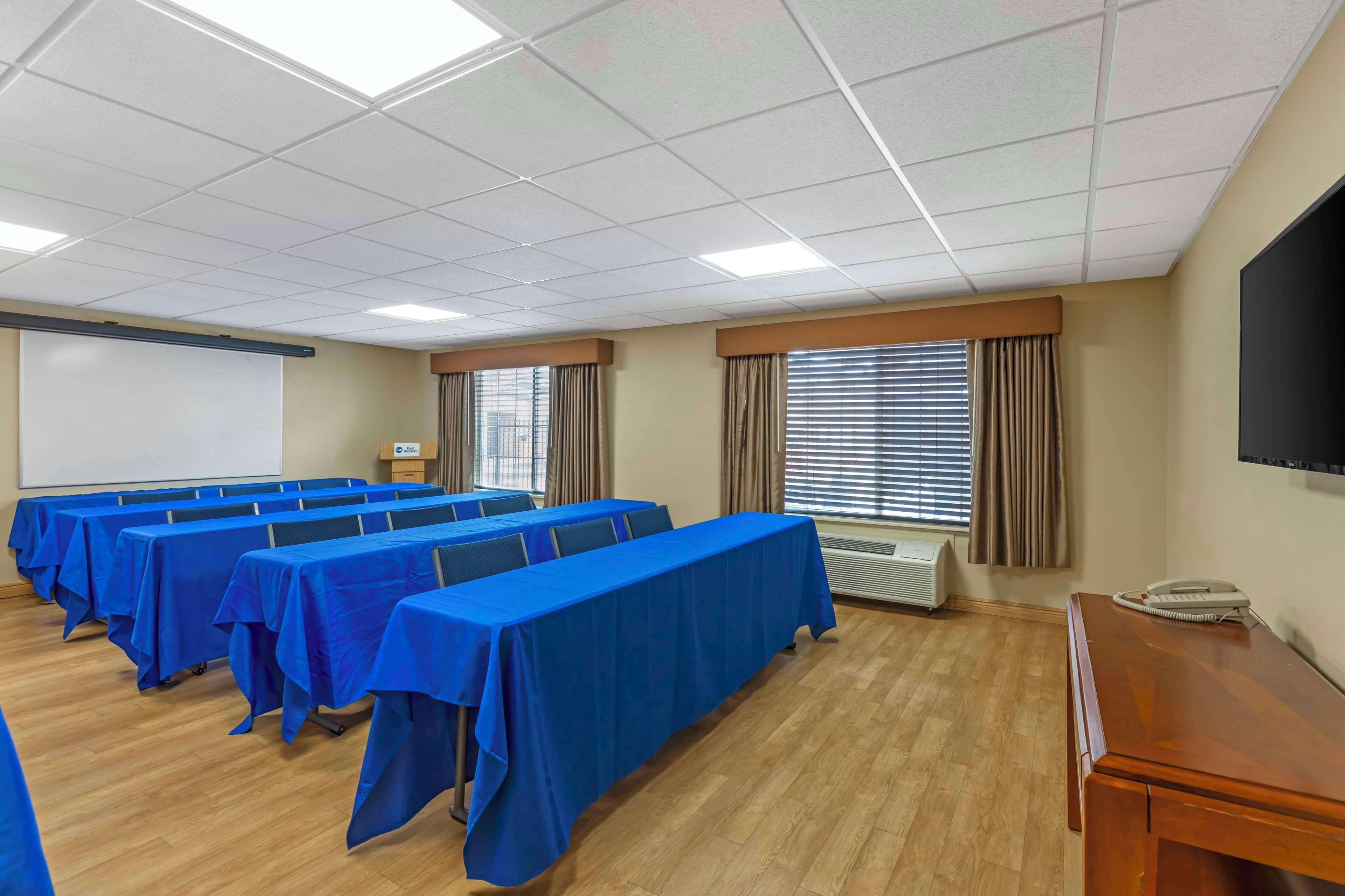 Une salle de conférence avec plusieurs tables recouvertes de nappes bleues, un écran de projection et un grand téléviseur à écran plat fixé au mur.