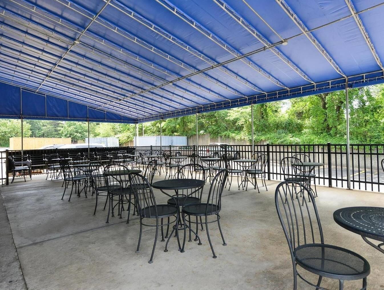 Un espace extérieur aménagé en terrasse, avec des tables et des chaises, recouvert d’un auvent bleu et entouré d’une clôture en métal noir.