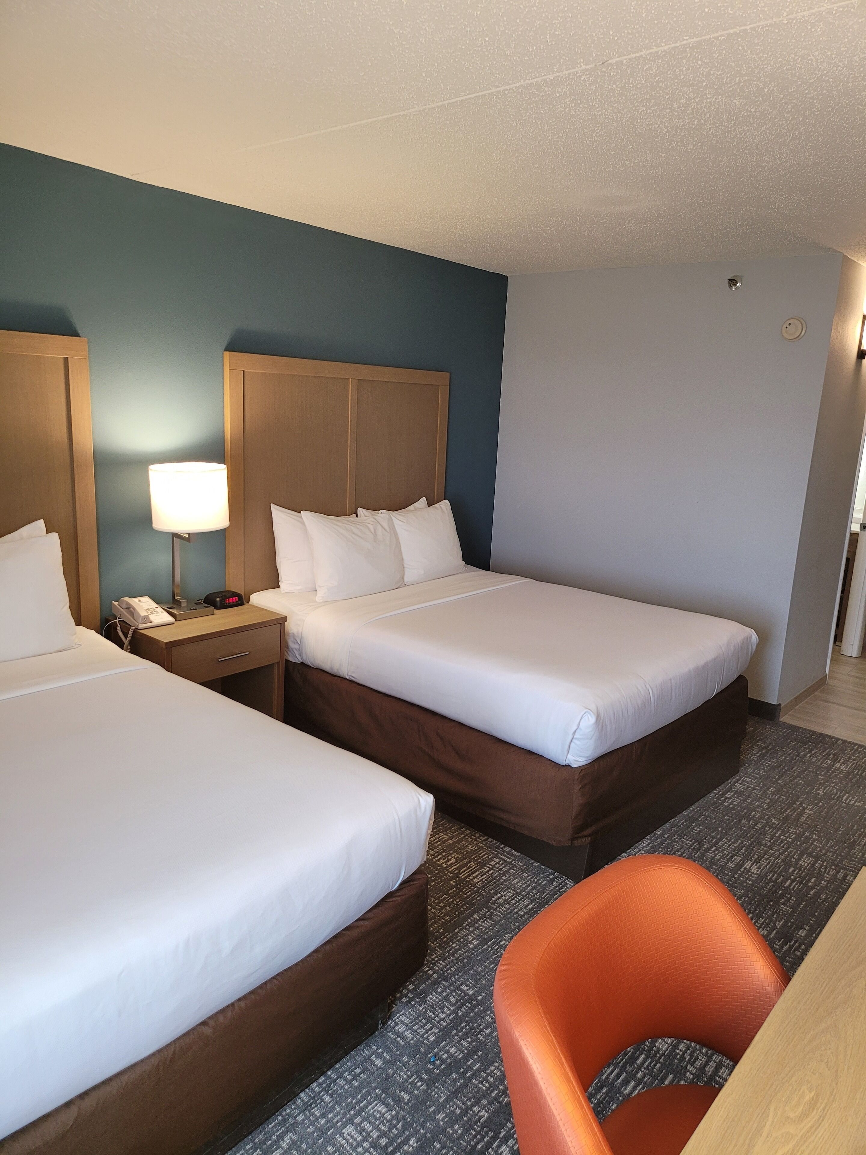 A hotel room with two beds, a nightstand with a lamp, a telephone, and an orange chair.
