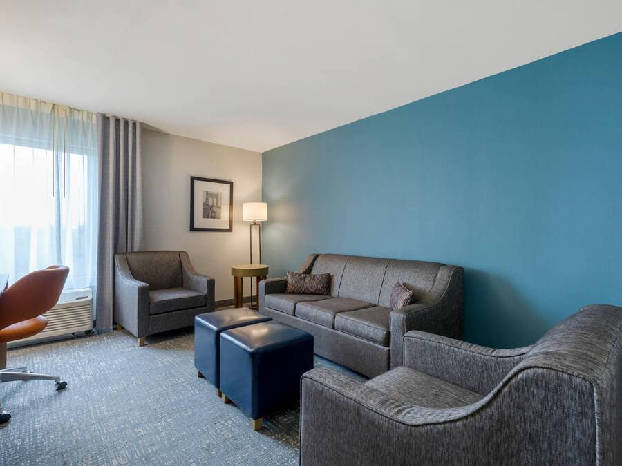 Una camera d'albergo con un divano grigio, una poltrona marrone, un poggiapiedi blu, una scrivania con una sedia, una lampada da terra e un quadro incorniciato appeso al muro.