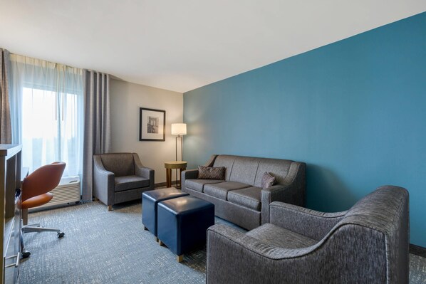 A hotel room with a grey sofa, a brown armchair, a blue ottoman, a desk with a chair, a standing lamp, and a framed picture on the wall. - Comfort Inn & Suites Alexandria West (Alexandria)