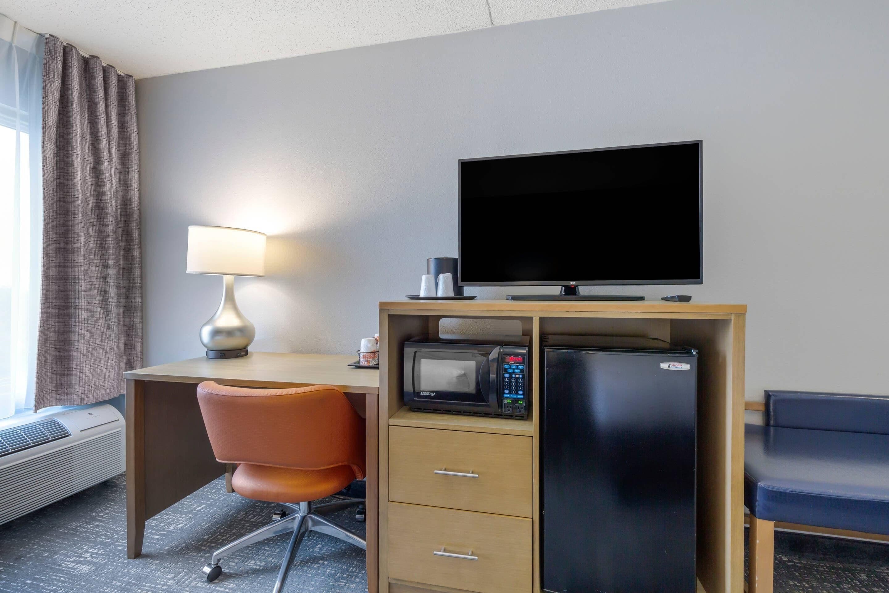 A hotel room with a desk, a television, a microwave, a refrigerator, an orange chair, and a lamp.