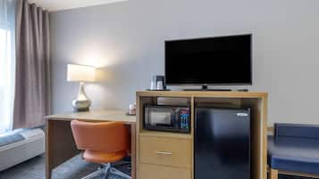 A hotel room with a desk, a television, a microwave, a refrigerator, an orange chair, and a lamp.