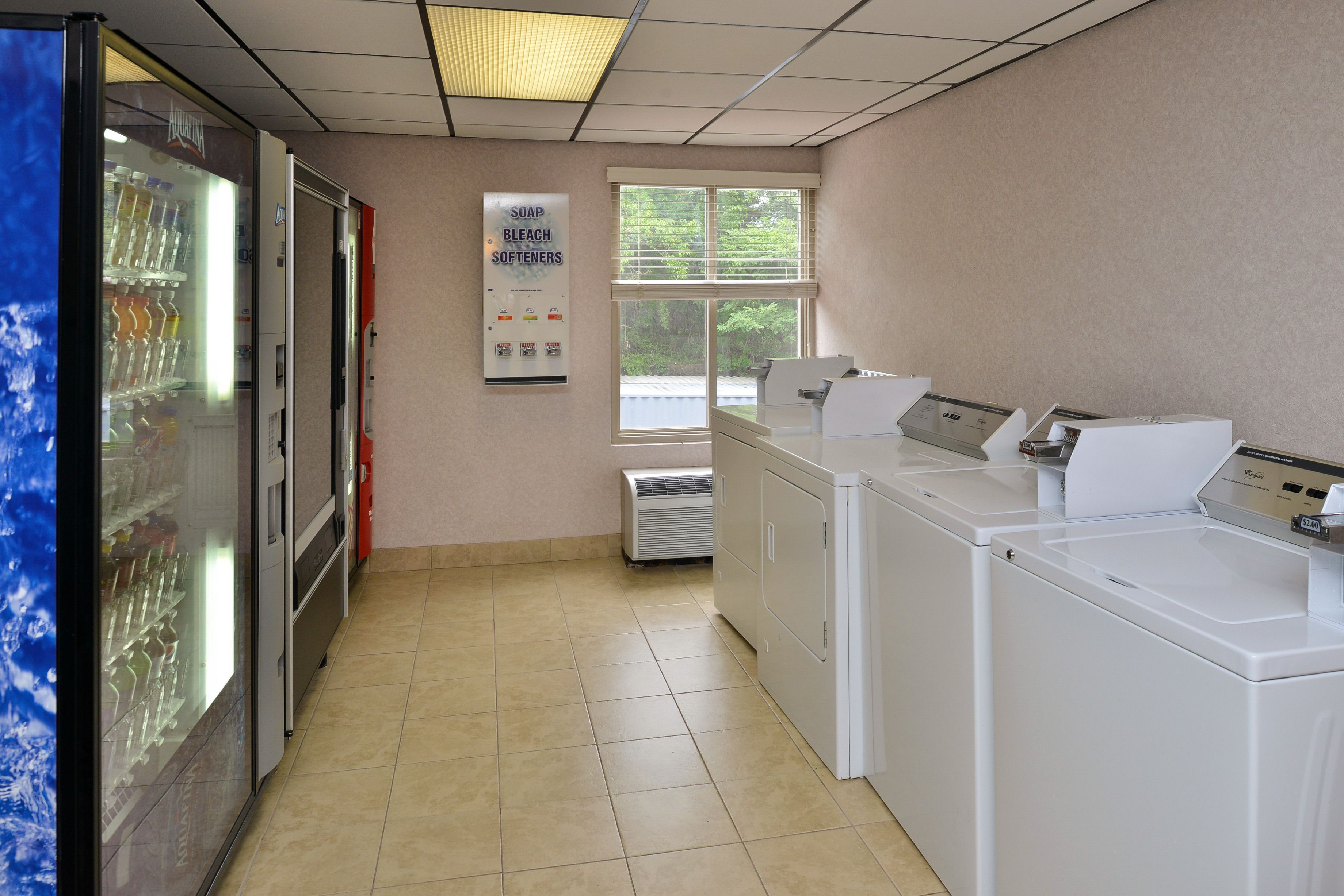 A laundry room with a vending machine, a window with blinds, and a sign about soap and bleach softeners.