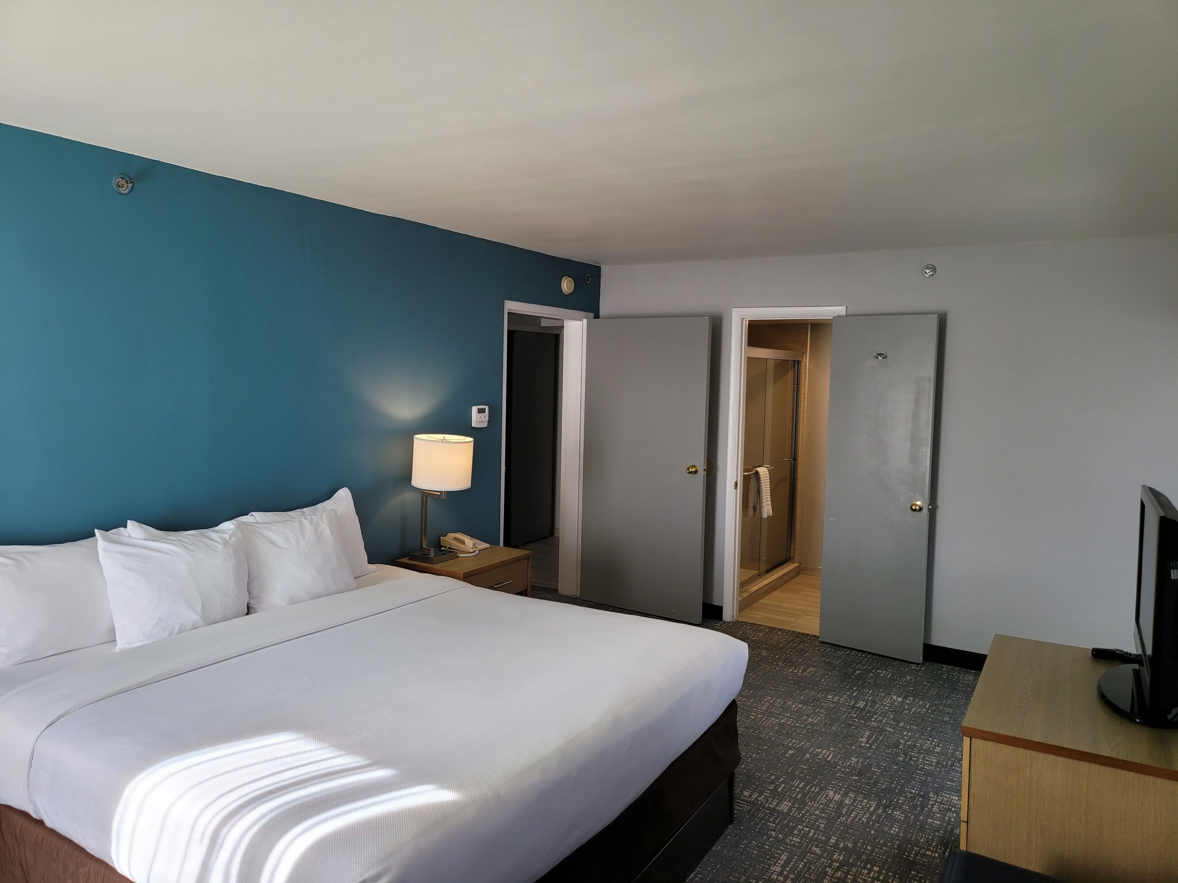 Habitación de hotel con cama, lámpara de noche, escritorio con televisor y zona de ducha.