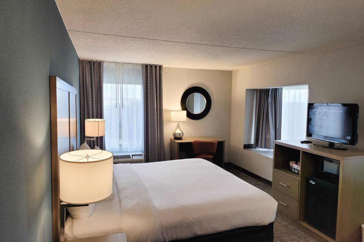 A hotel room with a bed, bedside table, lamp, TV, and a desk with a chair.