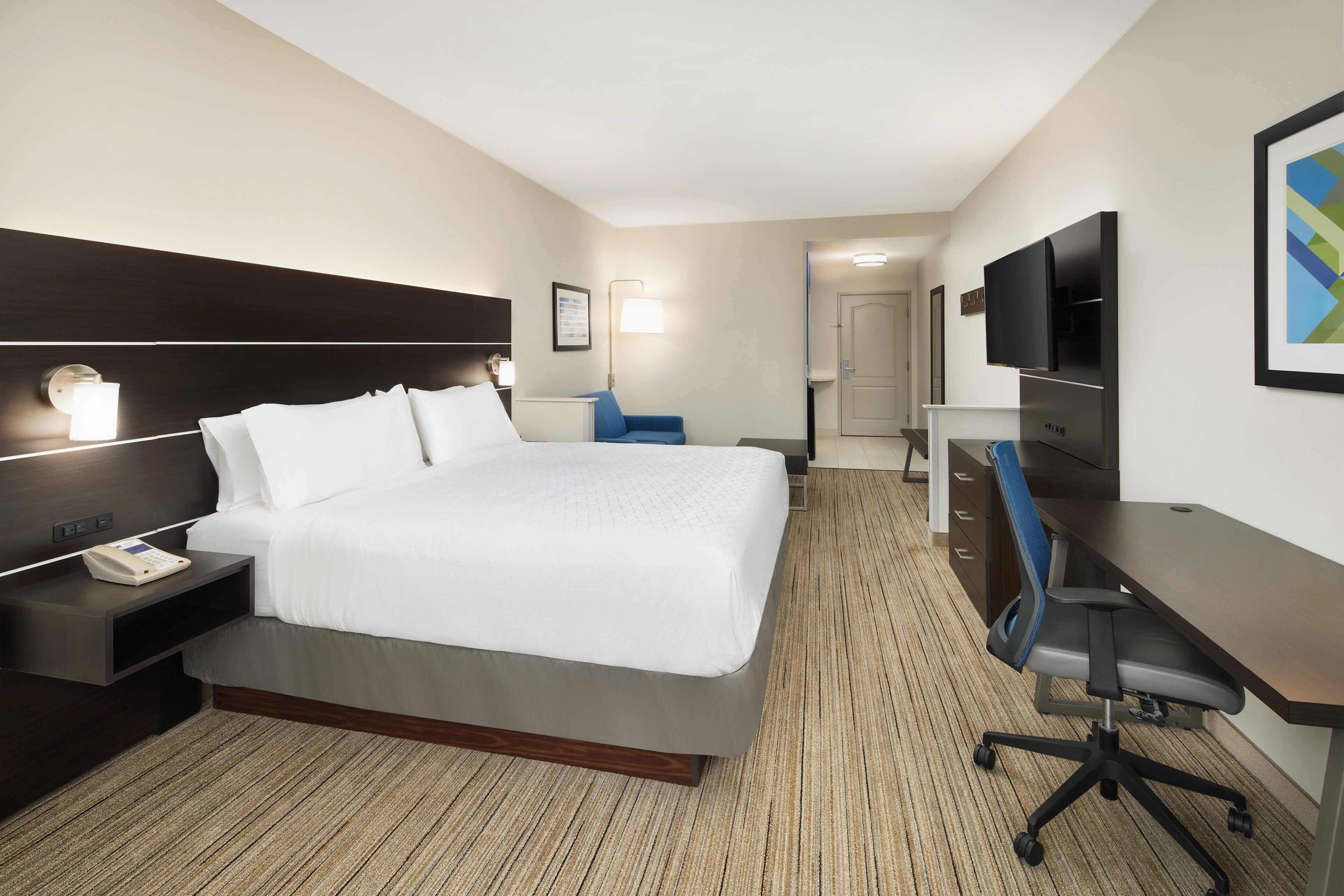 A hotel room with a bed, bedside table, lamp, desk, chair, and a television.