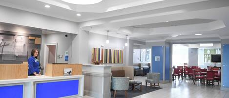 Zona de recepción de un hotel con un mostrador azul, una recepcionista y un área de descanso con sillas rojas.