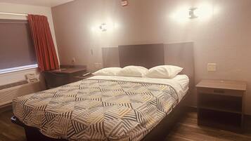 A hotel room with a bed, bedside tables, a desk, a chair, a window with curtains, and a wall-mounted light fixture.