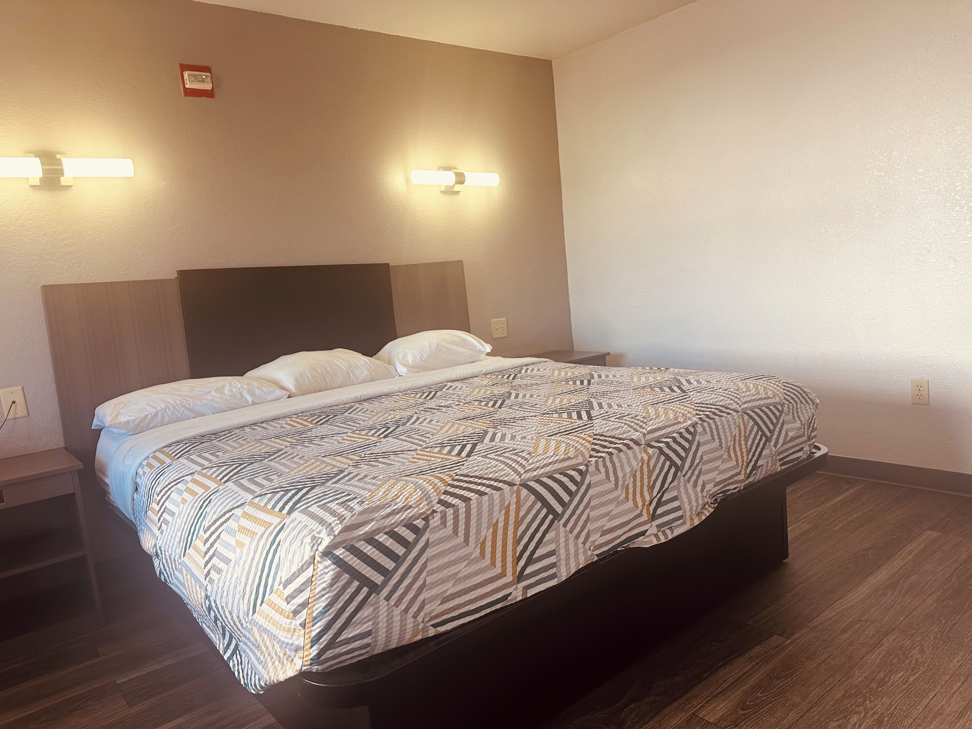 A hotel room with a bed, bedside tables, a headboard, wall-mounted lights, and a power outlet.