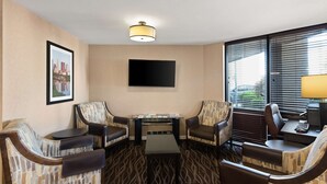 A hotel room with a sofa set, a small table, a flat-screen TV mounted on the wall, and a window with blinds. - Best Western Executive Inn (Grove City)