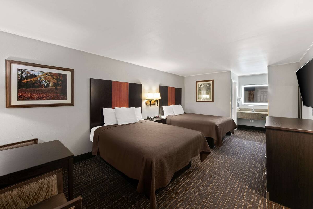 A hotel room with two beds, a desk, a chair, a lamp, and a TV.