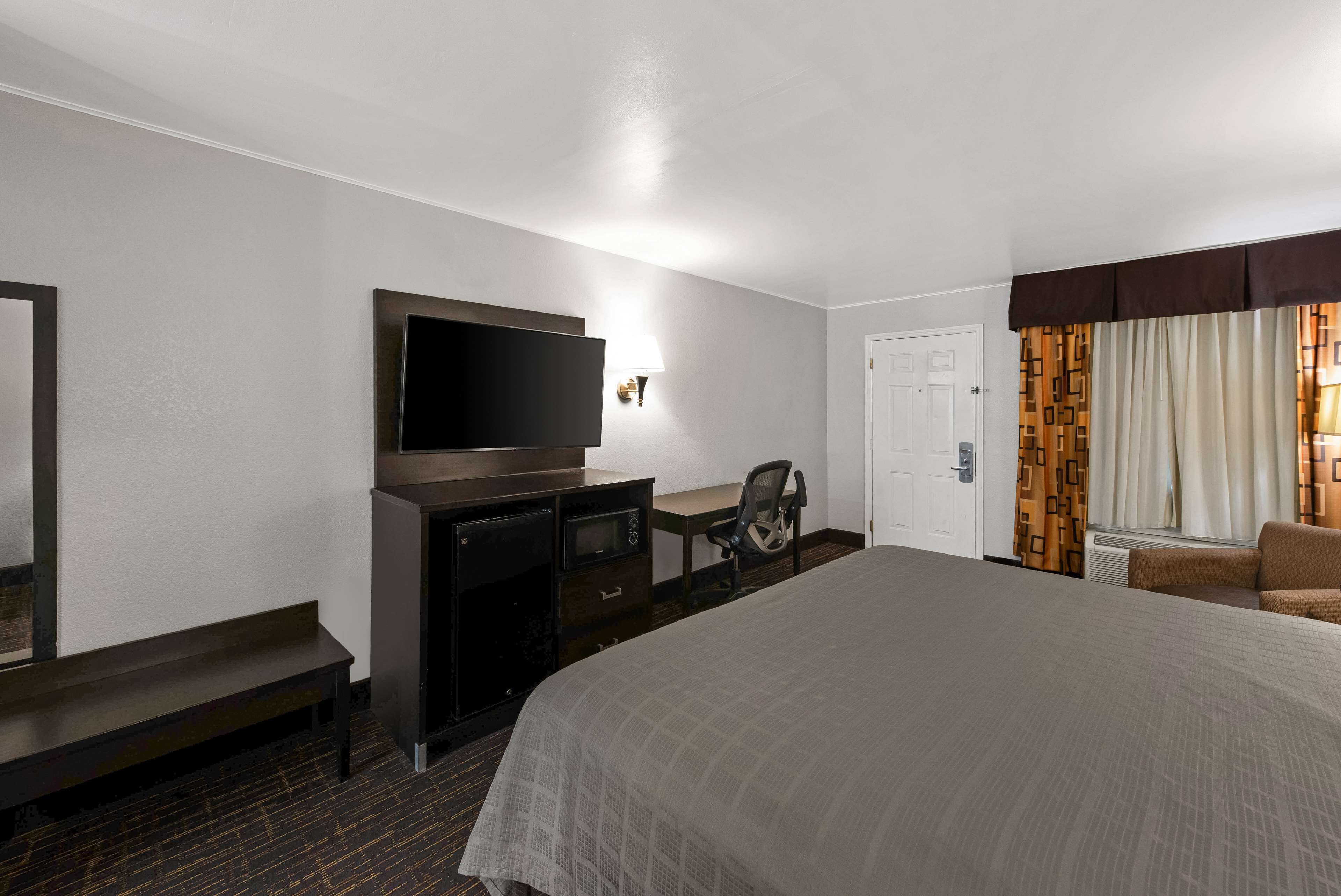 A hotel room with a large bed, a desk with a chair, a flat-screen TV, a dresser, and a window with patterned curtains.