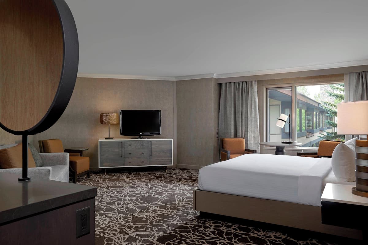 A hotel room with a large bed, a flat-screen TV on a cabinet, a desk with a lamp, and a view of the outdoors through a window.