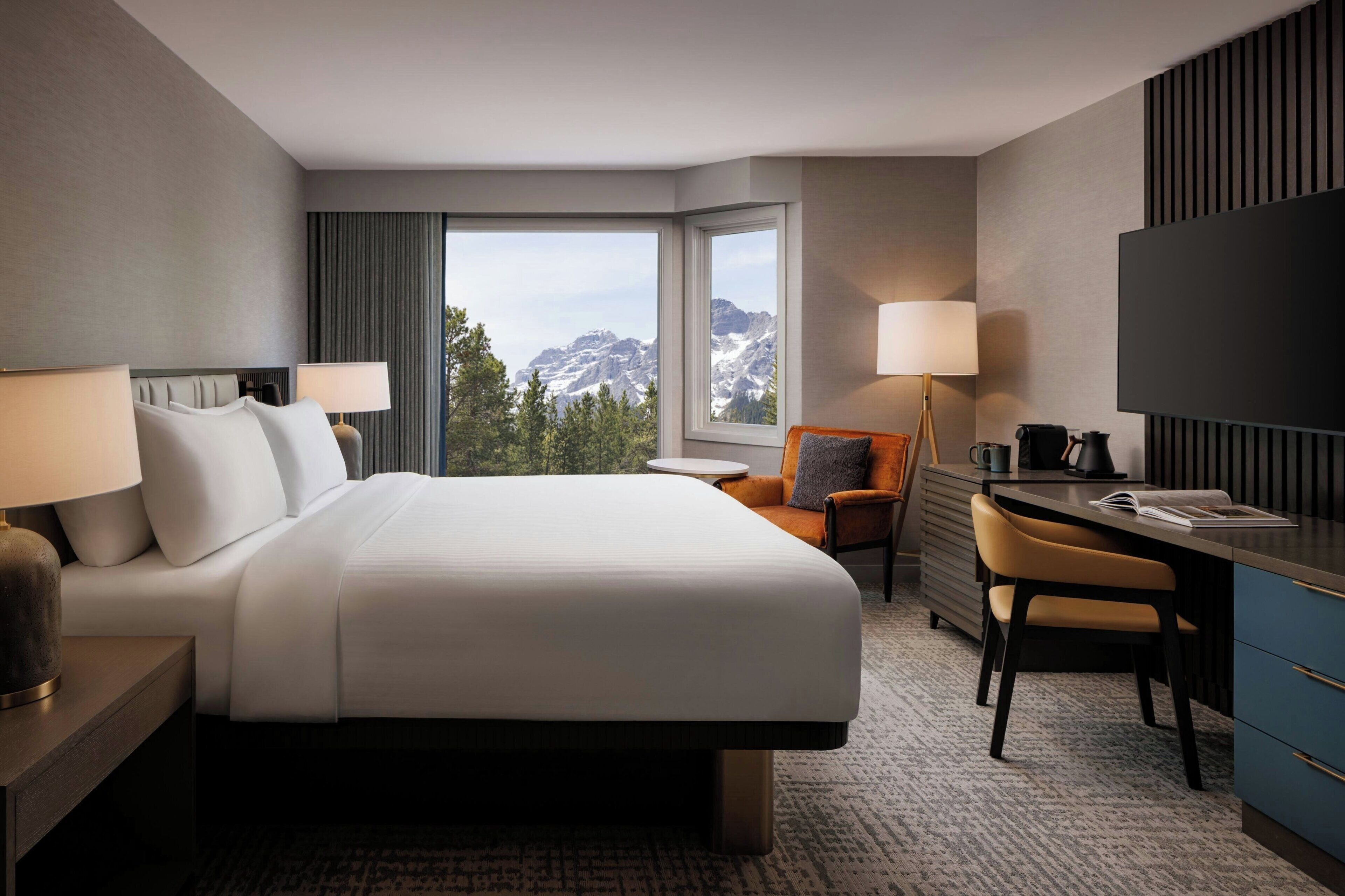 Habitación de hotel con una cama grande, un escritorio, una silla, un televisor y vistas a las montañas.
