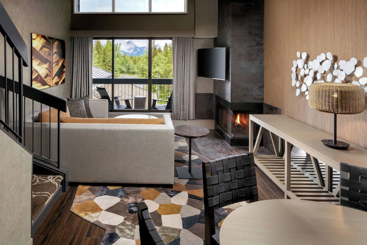 A modern living room with a sofa, a fireplace, a TV, and a balcony with a view of mountains.