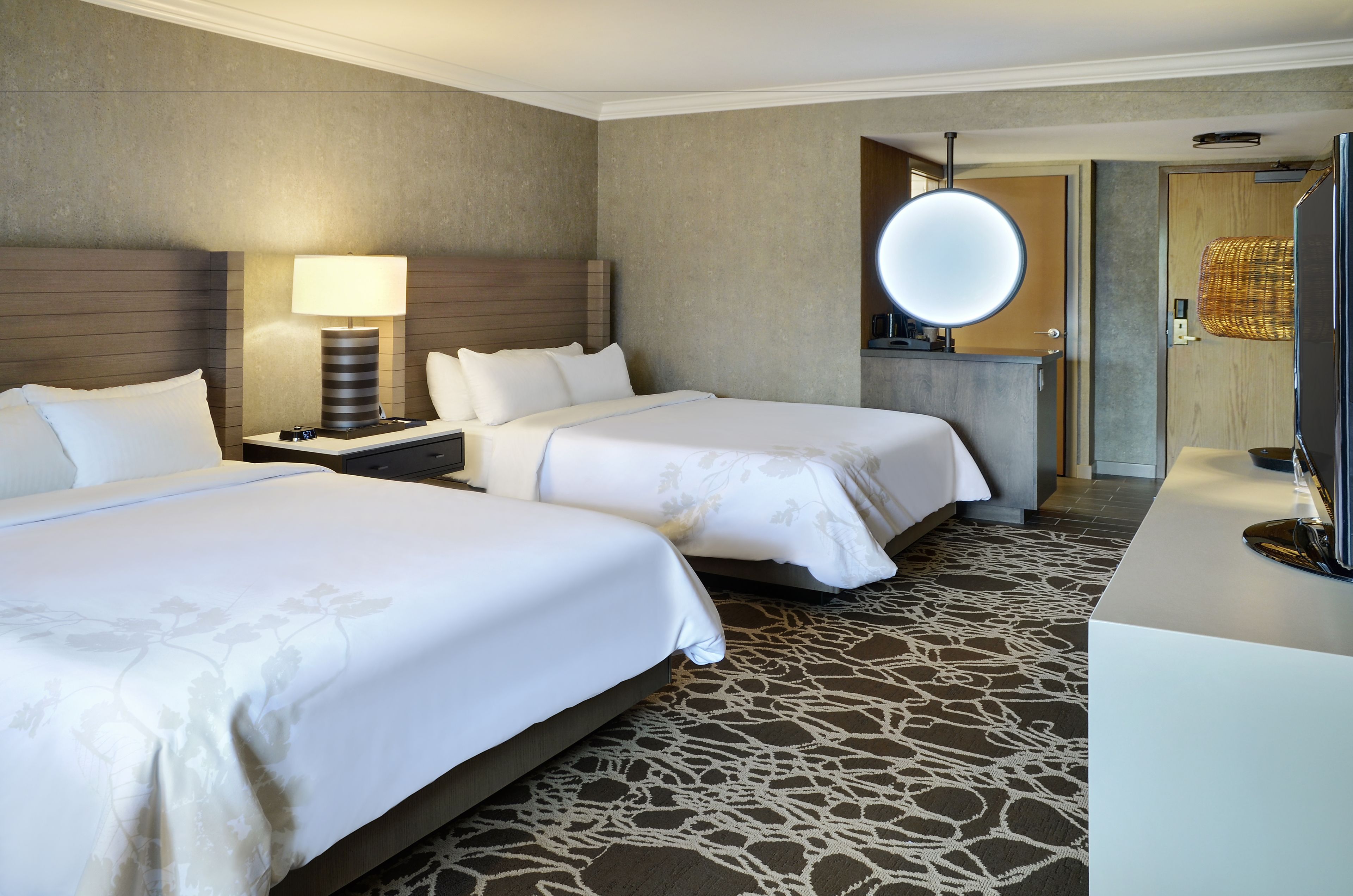 A hotel room with two beds, a large mirror, a nightstand, a lamp, and a modern bathroom with a large mirror.