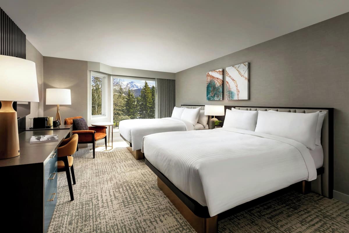 A hotel room with two beds, a desk with a lamp, a chair, and a view of mountains through the window.