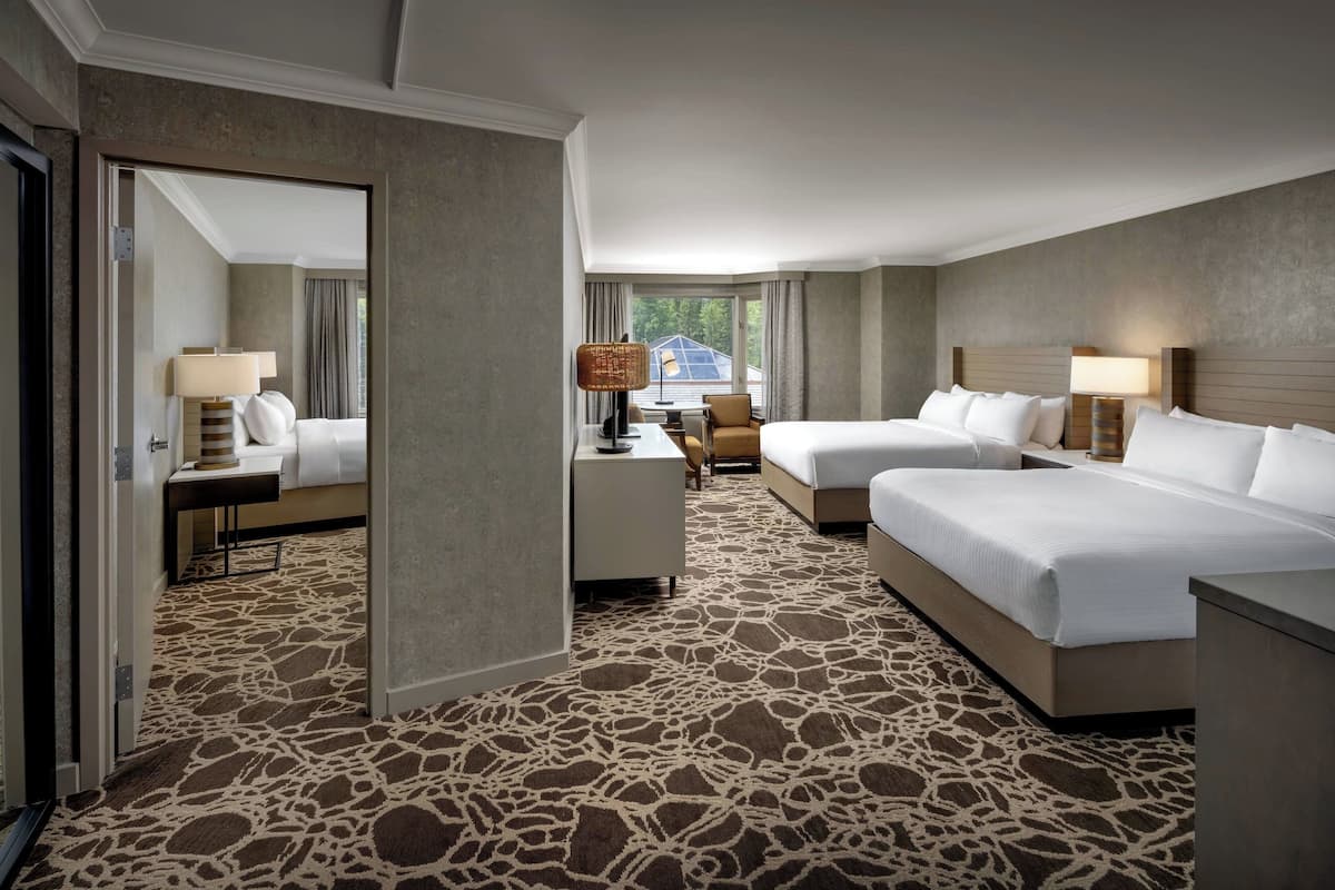 A hotel room with two beds, a desk, and a chair. The room has a patterned carpet and a view of the outdoors.