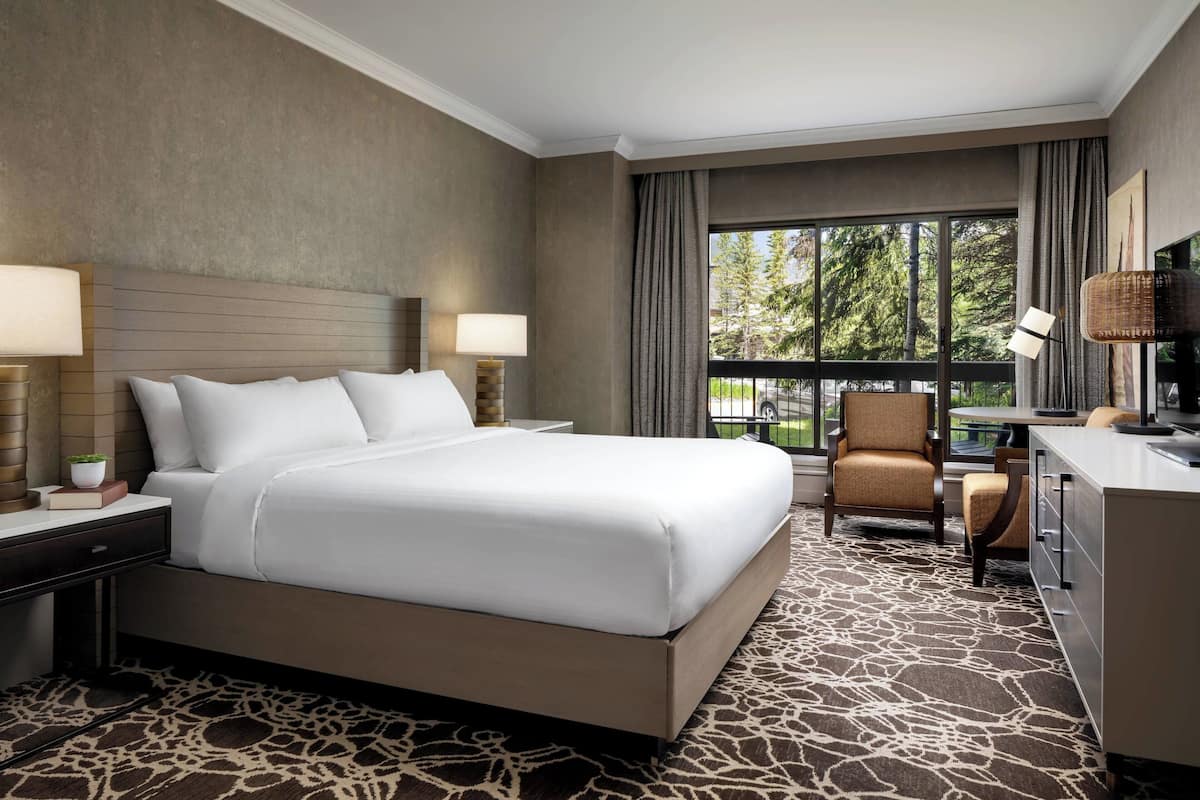 A hotel room with a large bed, two bedside tables with lamps, a desk with a chair, and a view of trees through the window.