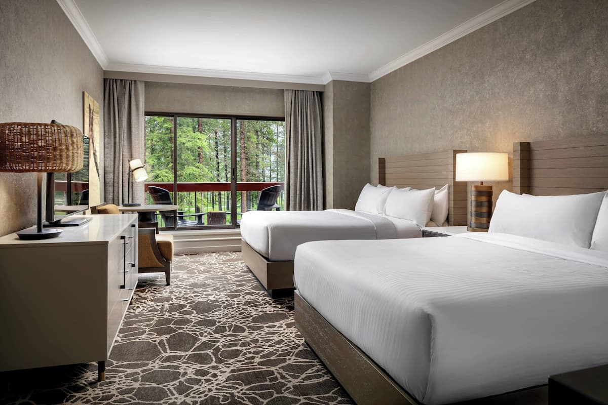 A hotel room with two beds, a desk, a chair, a lamp, and a view of trees through large windows.
