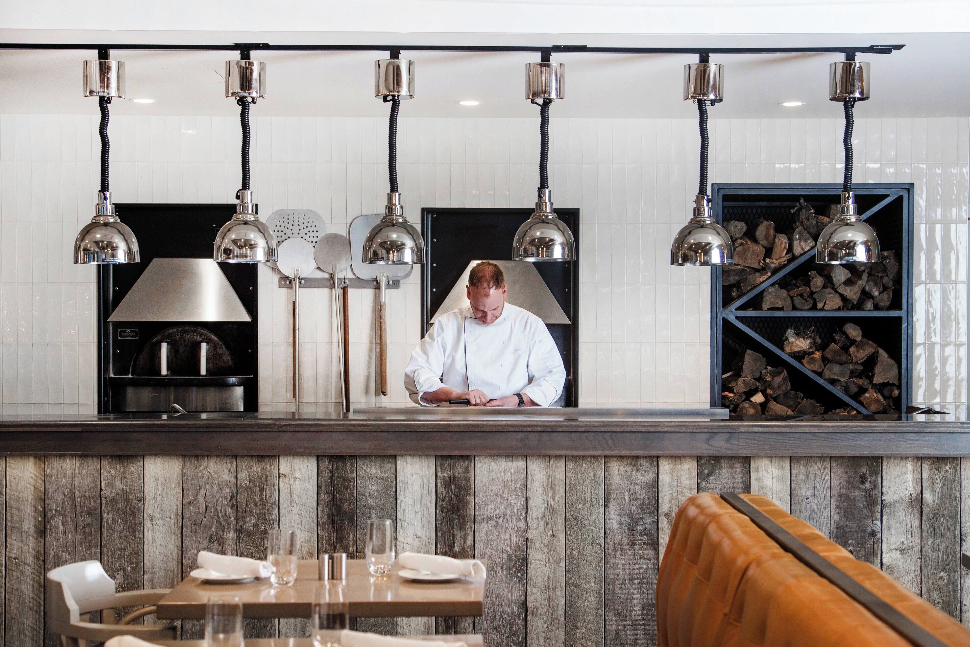 Un chef travaille derrière un comptoir dans un restaurant dont le mur est en bois, où pendent des lampes décoratives, et qui dispose d’un espace salon avec un banc en bois.