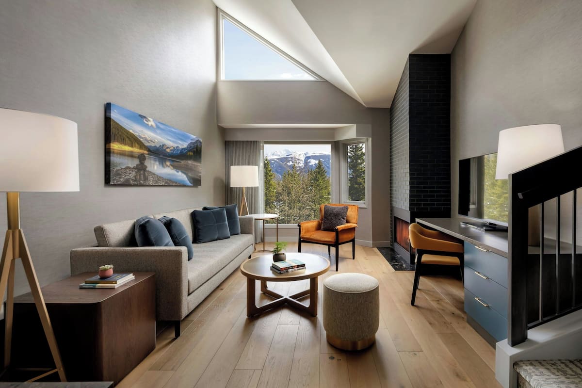 A modern living room with a sofa, coffee table, and armchairs, a painting on the wall, and a view of mountains through large windows.