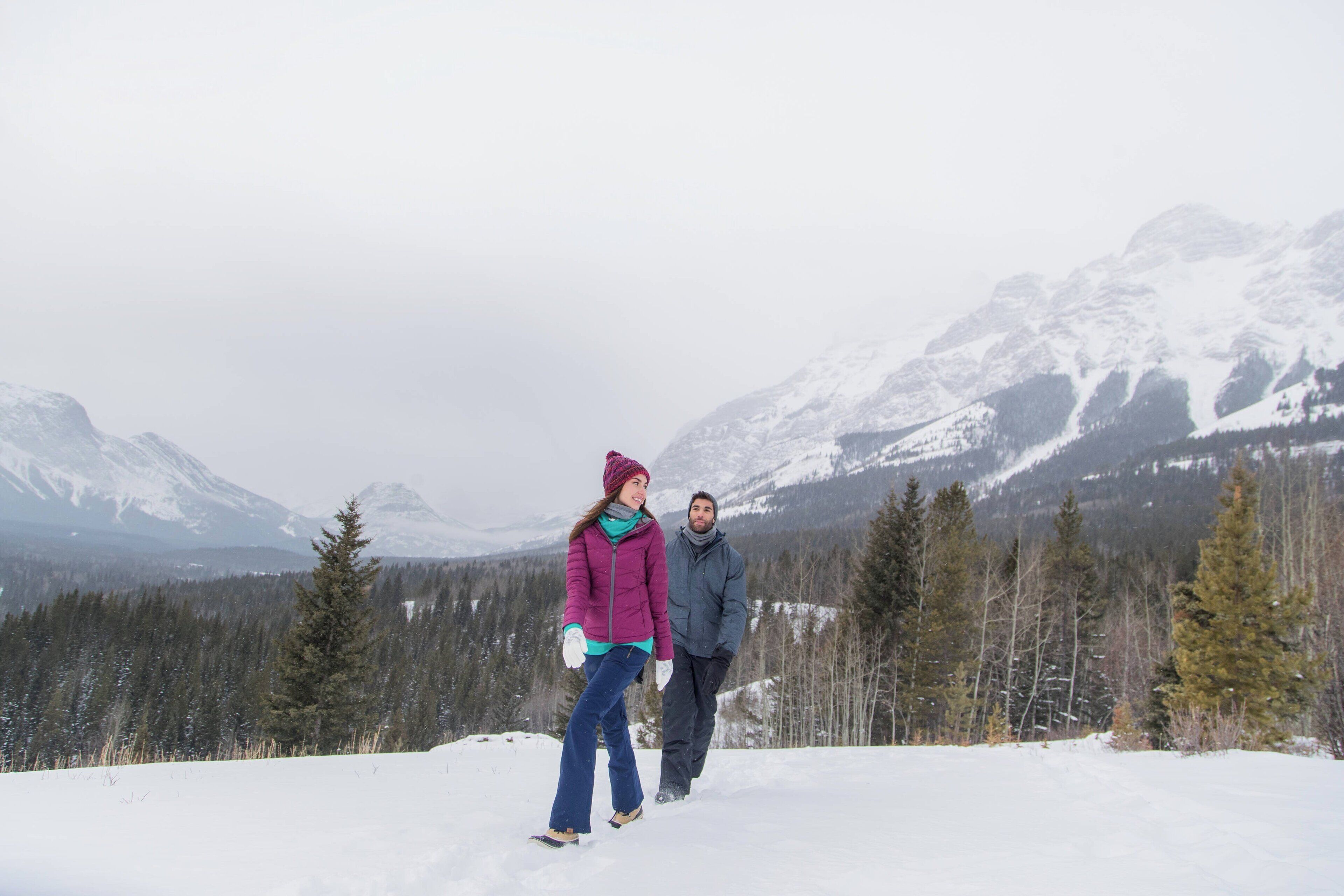 A couple walking on a snow-covered path with a mountainous backdrop.