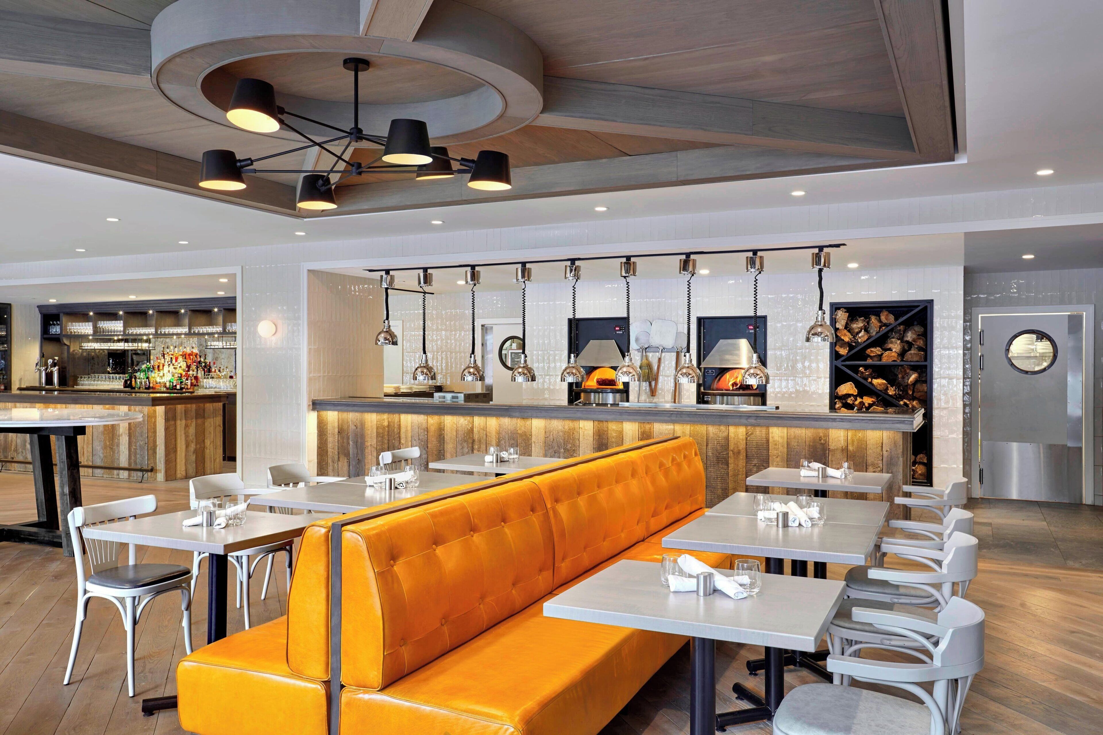 Ein modernes Restaurant mit einer zentralen orangefarbenen Essecke, weißen Tischen und hängenden Pendelleuchten.